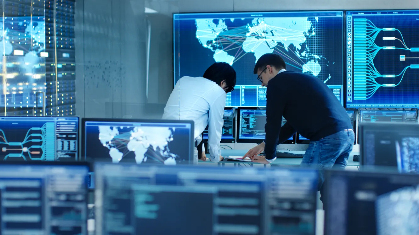Two AI engineers working at data center terminals with global network map and system dashboards on large screens, representing generative AI and global pharma talent