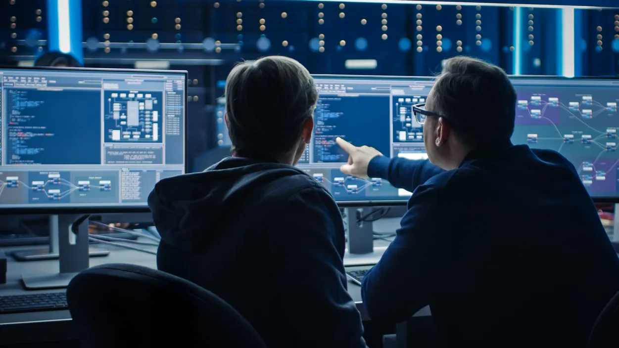 Two engineers reviewing complex AI system architecture on dual monitors in a dark server operations room, representing drug discovery AI development
